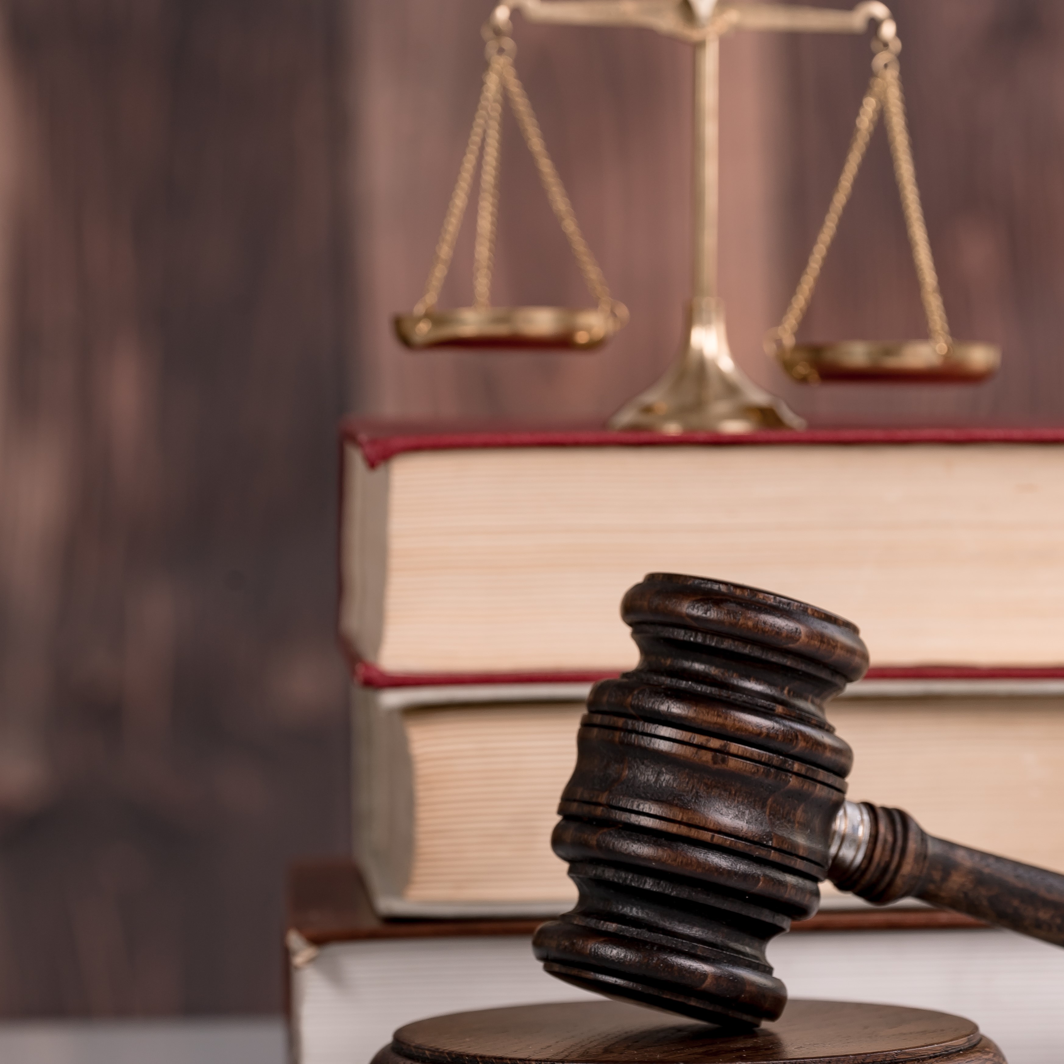 A wooden gavel rests on a round base, in front of stacked law books with red bindings. Behind the gavel is a traditional brass balance scale, symbolizing justice. The background features a blurred wood grain texture.