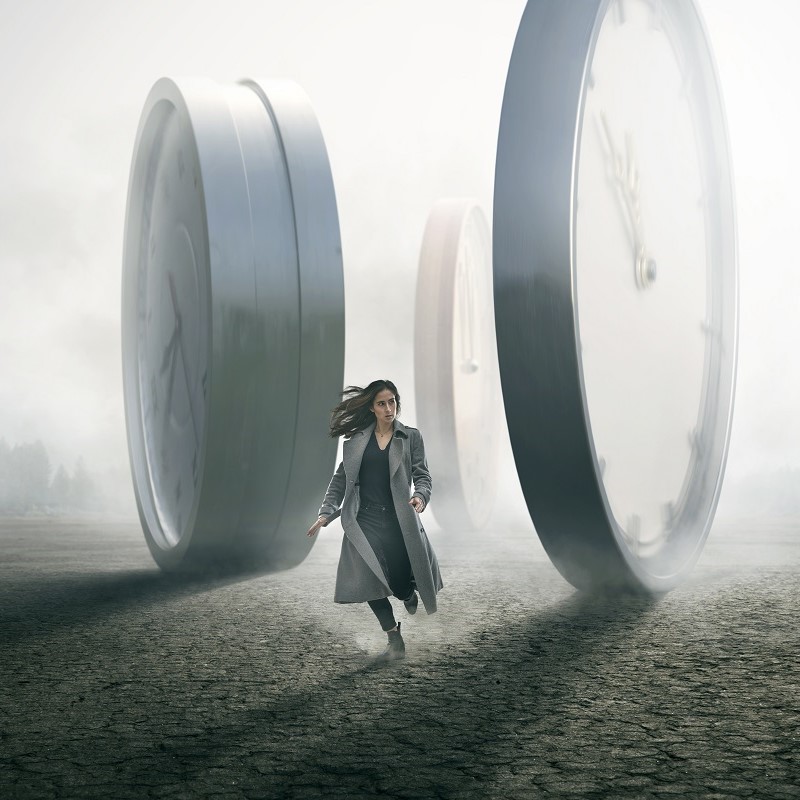 A woman in a gray coat runs across a cracked ground, flanked by large, surreal clocks looming in the mist. The scene evokes a sense of urgency and the passage of time, highlighting the contrast between her movement and the stationary clocks.