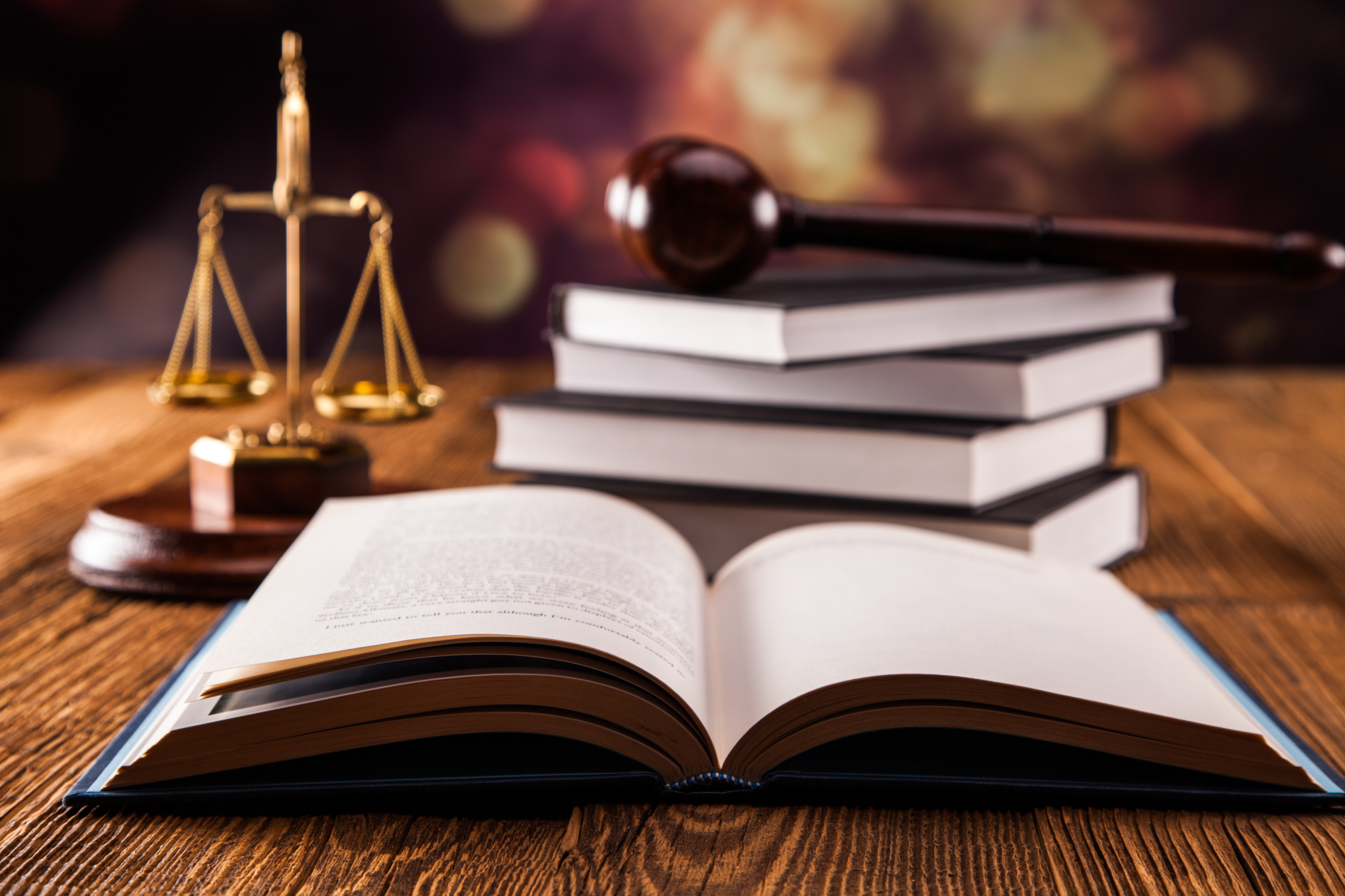 An open law book sits on a wooden table, with several stacked books beside it. A golden scale of justice is prominently displayed in the background, along with a gavel placed diagonally. The setting suggests a legal or courtroom theme.