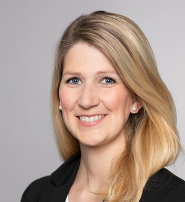A professional woman with long, blonde hair and blue eyes smiles confidently. She wears a black blazer and has pearl earrings. The background is a light gray, enhancing her approachable demeanor.