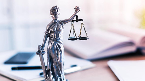 Kartellschadensersatz Eine silberne Statue der Justitia mit Augenbinde, die eine Waage in der einen Hand und ein Schwert in der anderen hält. Sie symbolisiert Gerechtigkeit und Rechtsprechung. Der Hintergrund zeigt verschwommene Notizen und Schreibutensilien.