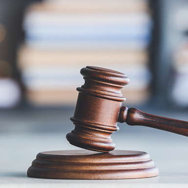 Ein hölzerner Richterhammer liegt auf einem runden Podest. Der Hintergrund ist unscharf, zeigt jedoch Stapel von Akten oder Büchern. Der Hammer symbolisiert rechtliche Entscheidungen und die Autorität des Gerichts.