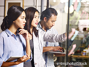 Three individuals work collaboratively in a modern office setting. One woman holds a tablet, while another woman observes intently. A man sits nearby, focused on typing on a laptop. Their expressions convey concentration and teamwork.