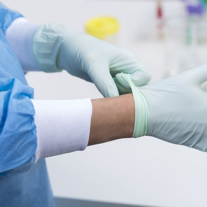 A close-up of a hand wearing a light green latex glove, as it pulls the glove onto the wrist. The hand is partially covered by a blue surgical gown and is positioned near a laboratory setting.